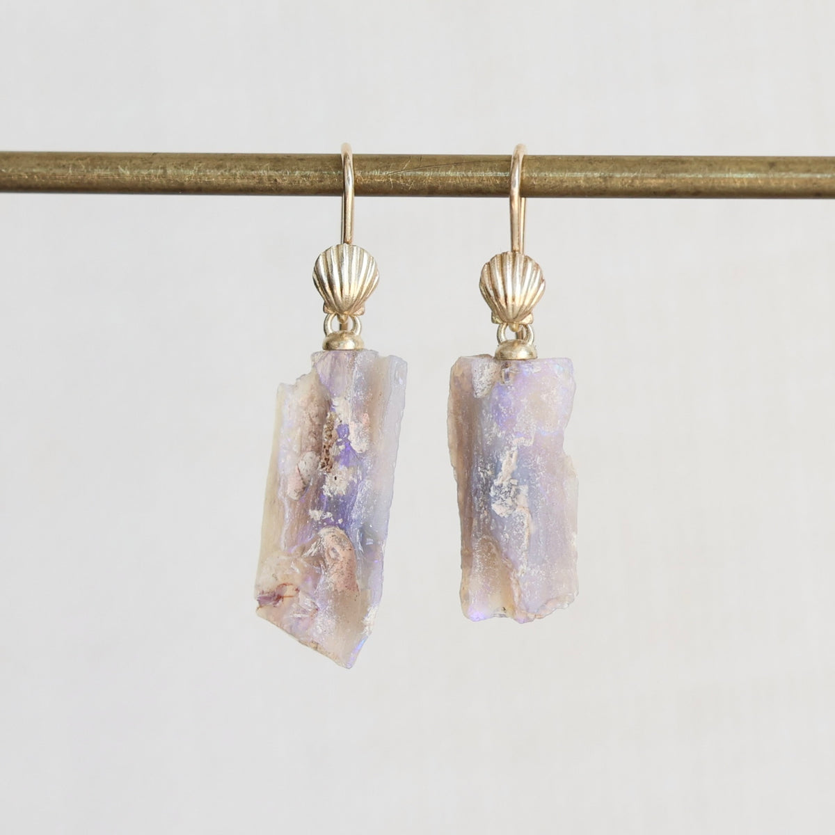A pair of gold drop earrings with scallop-shaped gold tops and irregular, raw opal-rectangular pendants showing lavender, purple, and cream iridescence, hanging from a gold rod against a light gray background.