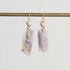 A pair of gold drop earrings with scallop-shaped gold tops and irregular, raw opal-rectangular pendants showing lavender, purple, and cream iridescence, hanging from a gold rod against a light gray background.