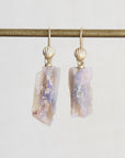 A pair of gold drop earrings with scallop-shaped gold tops and irregular, raw opal-rectangular pendants showing lavender, purple, and cream iridescence, hanging from a gold rod against a light gray background.
