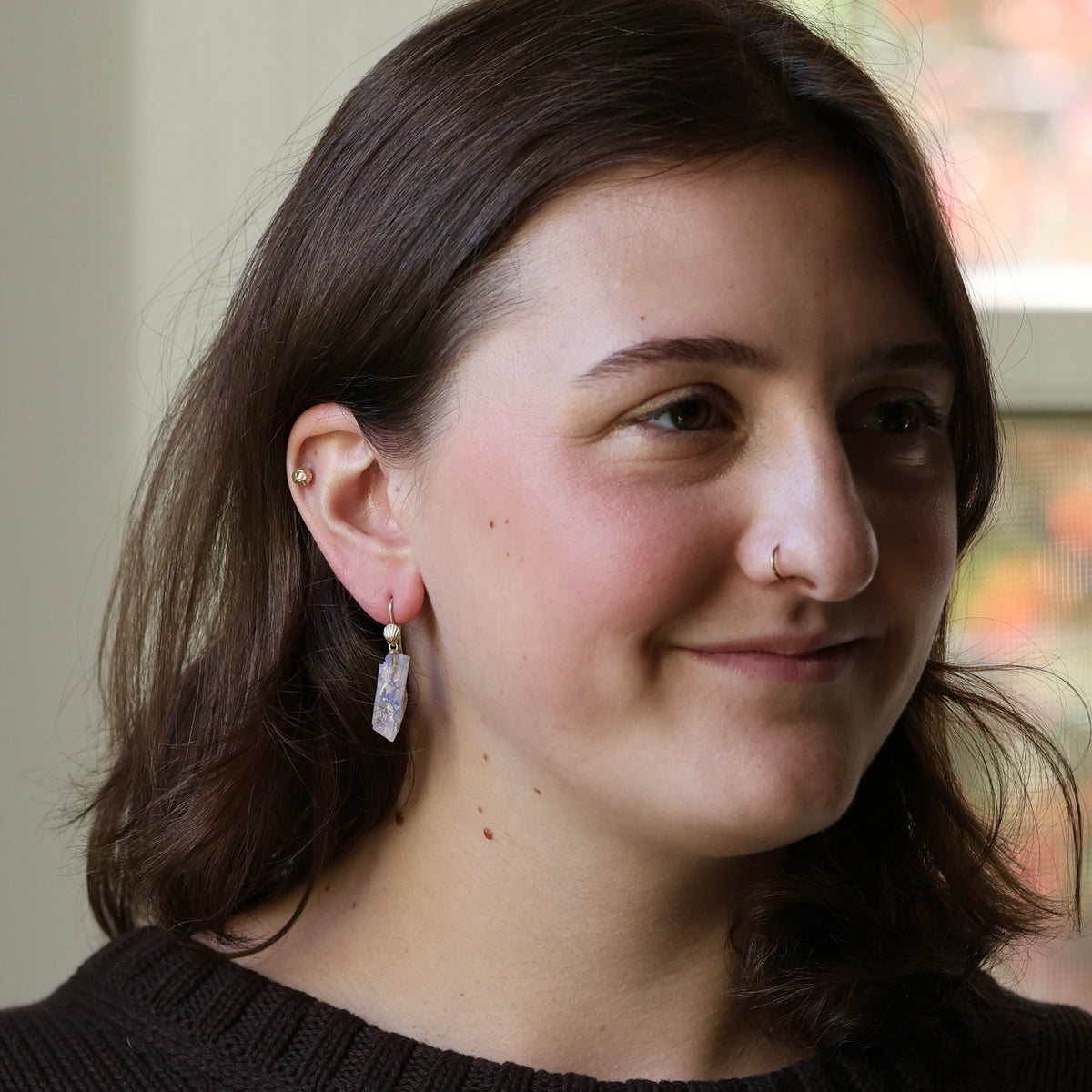 A close-up portrait of a woman with brown hair wearing a dangling rectangular crystal-like earring with a gold-toned setting and clear stone, shown in a softly lit indoor setting.