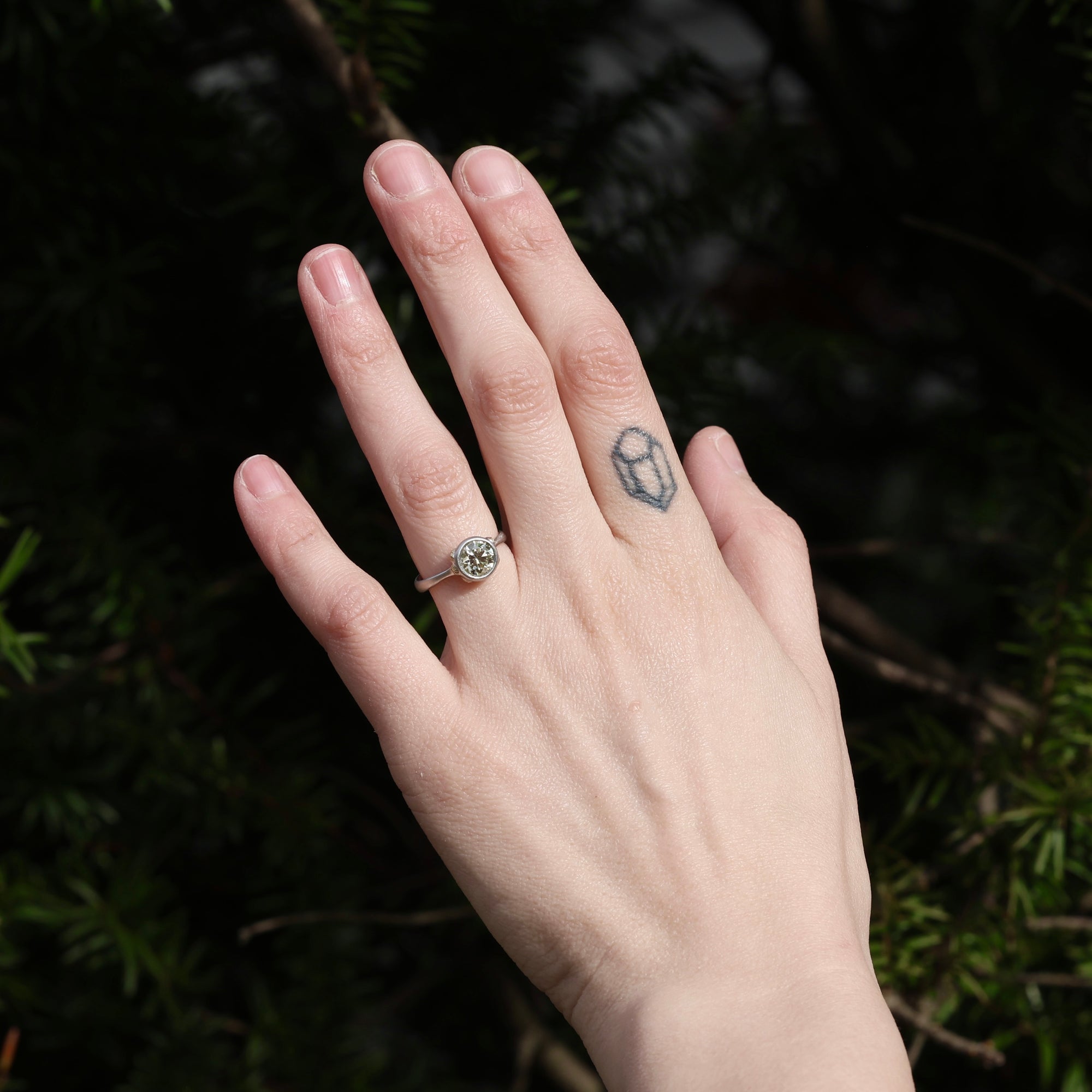 Hand with a ring and tattoo against a dark green leafy background