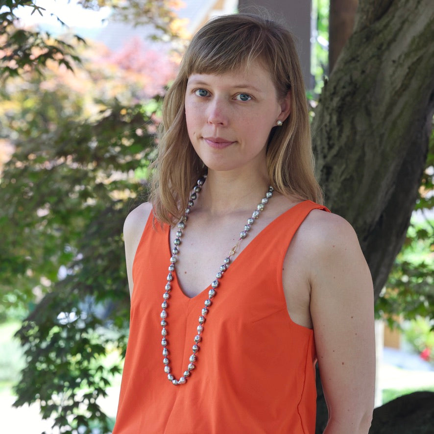 Woman wearing an orange sleeveless top with a long necklace, standing outdoors with trees in the background.