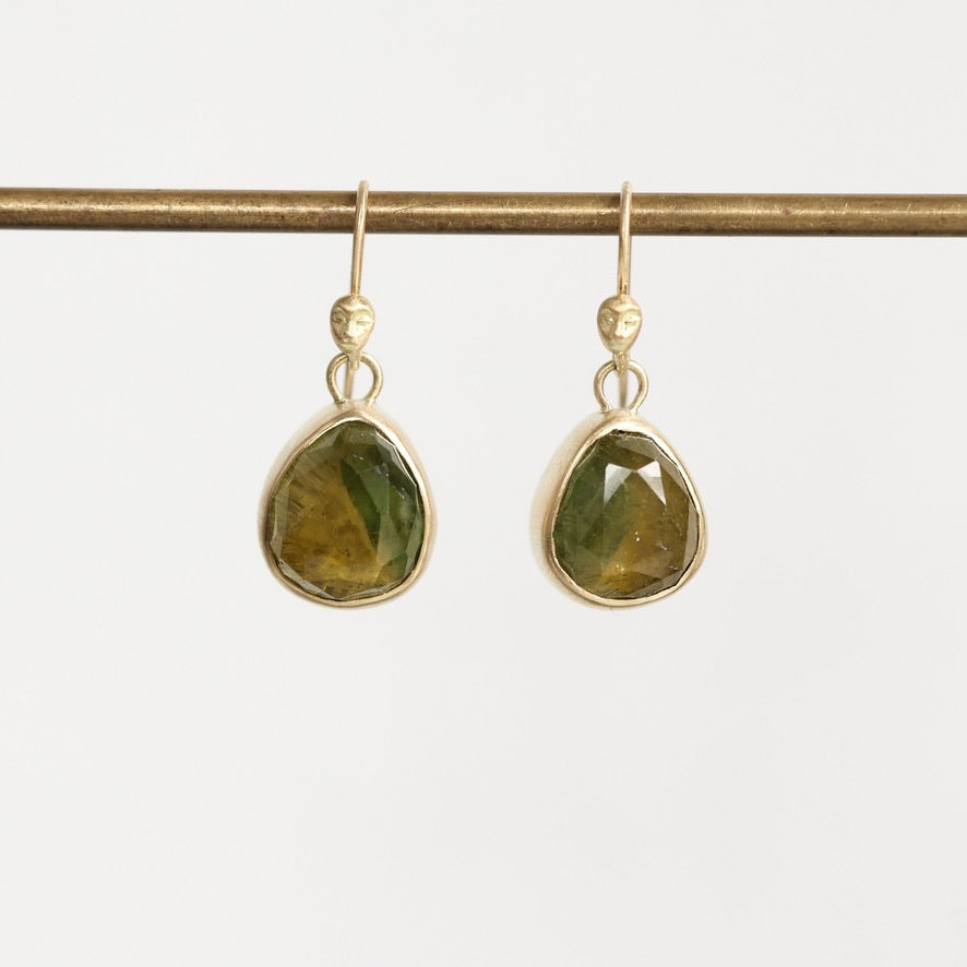 A pair of gold drop earrings with faceted green teardrop-shaped gemstones, hanging from a gold rod against a light gray background.