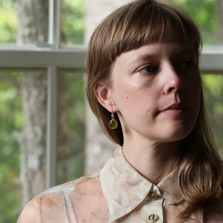 A close-up portrait of a woman with light brown hair and bangs, wearing a patterned cream blouse and a small gold hoop earring with a green teardrop charm, set against a softly blurred window background.