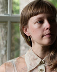 A close-up portrait of a woman with light brown hair and bangs, wearing a patterned cream blouse and a small gold hoop earring with a green teardrop charm, set against a softly blurred window background.