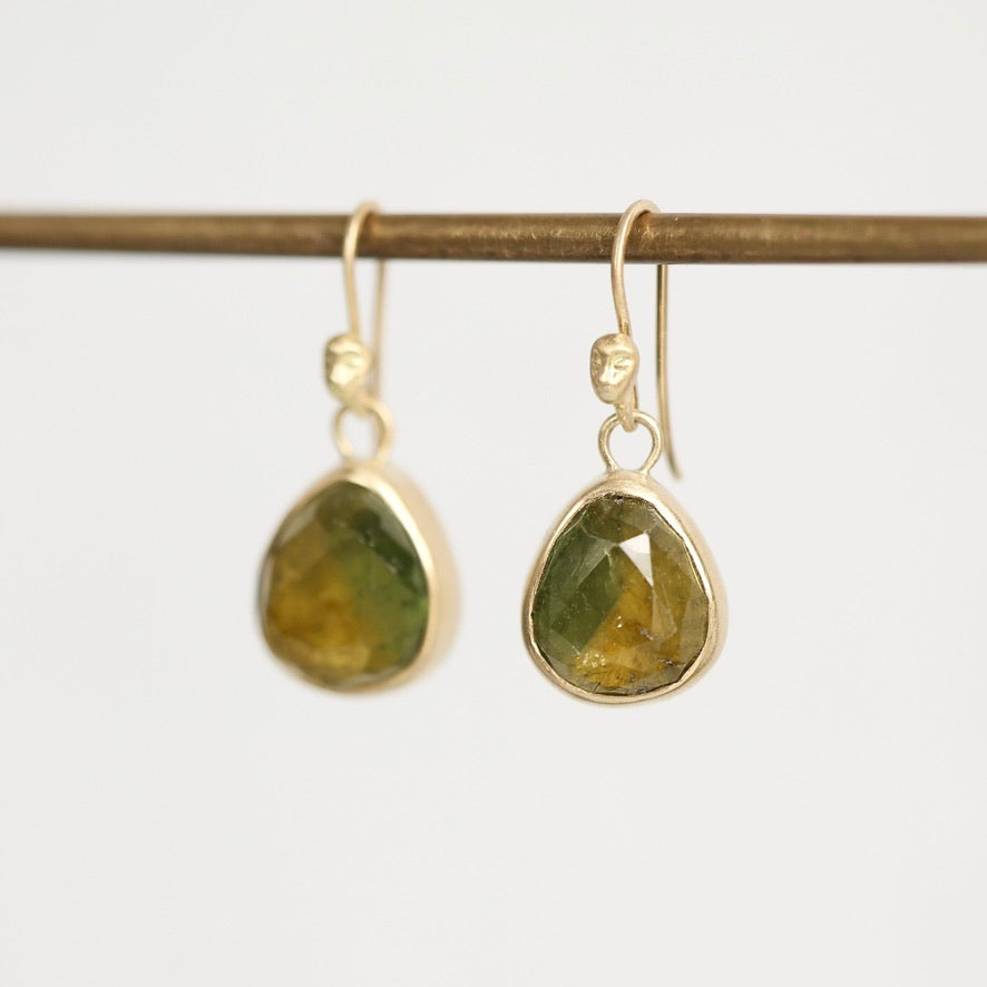 A pair of gold drop earrings with faceted green gemstone pendants, hanging from a thin horizontal gold rod against a plain light gray background.