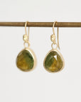 A pair of gold drop earrings with faceted green gemstone pendants, hanging from a thin horizontal gold rod against a plain light gray background.