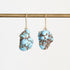 A pair of dangling earrings with irregular light blue turquoise-like stones set in gold-toned metal, hanging from a gold rod against a plain light background.