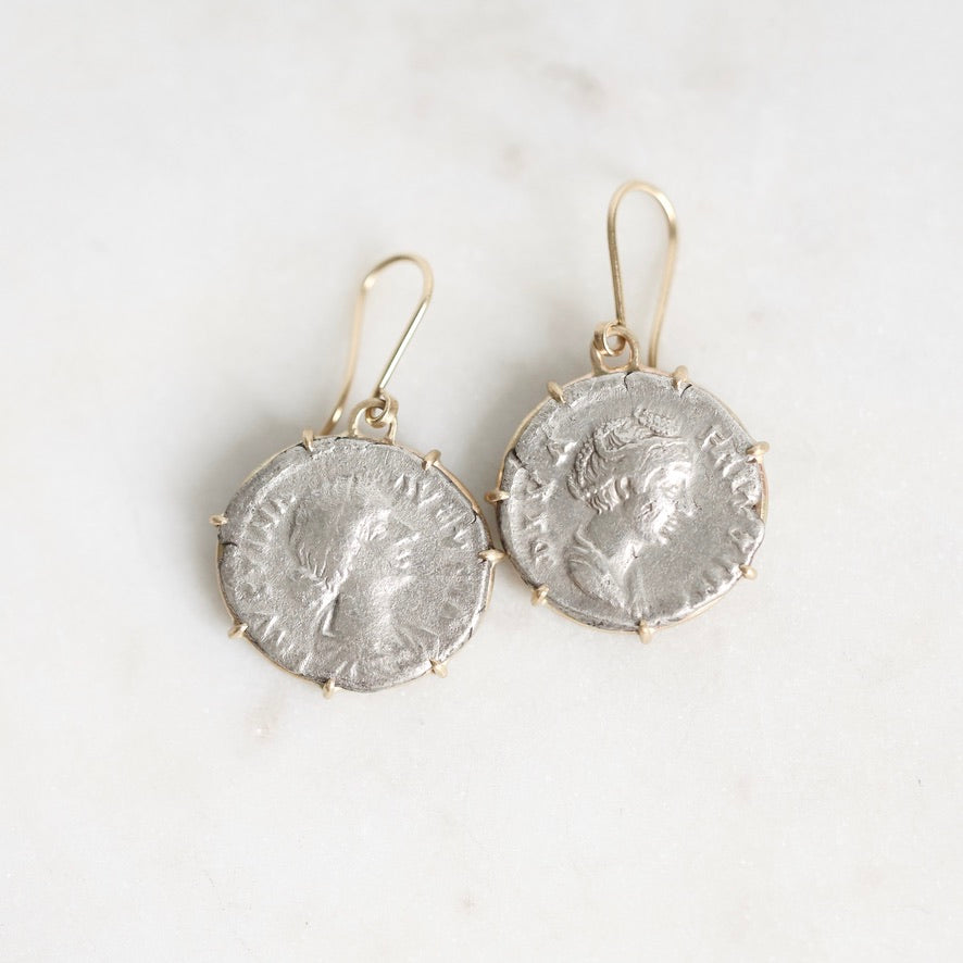 A pair of dangling earrings made from silver-toned ancient coin faces set in gold-toned metal hooks, displayed on a light marble surface.