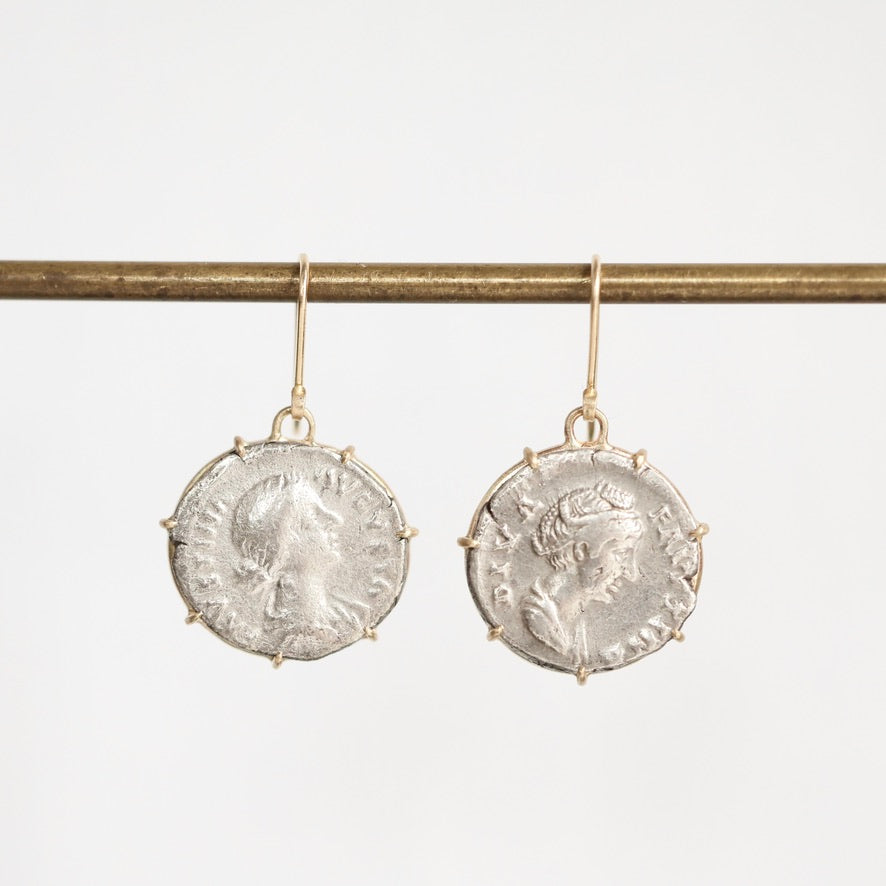 A pair of dangling earrings shaped like aged silver coins with gold-toned hooks, hanging from a horizontal gold rod against a plain light background.
