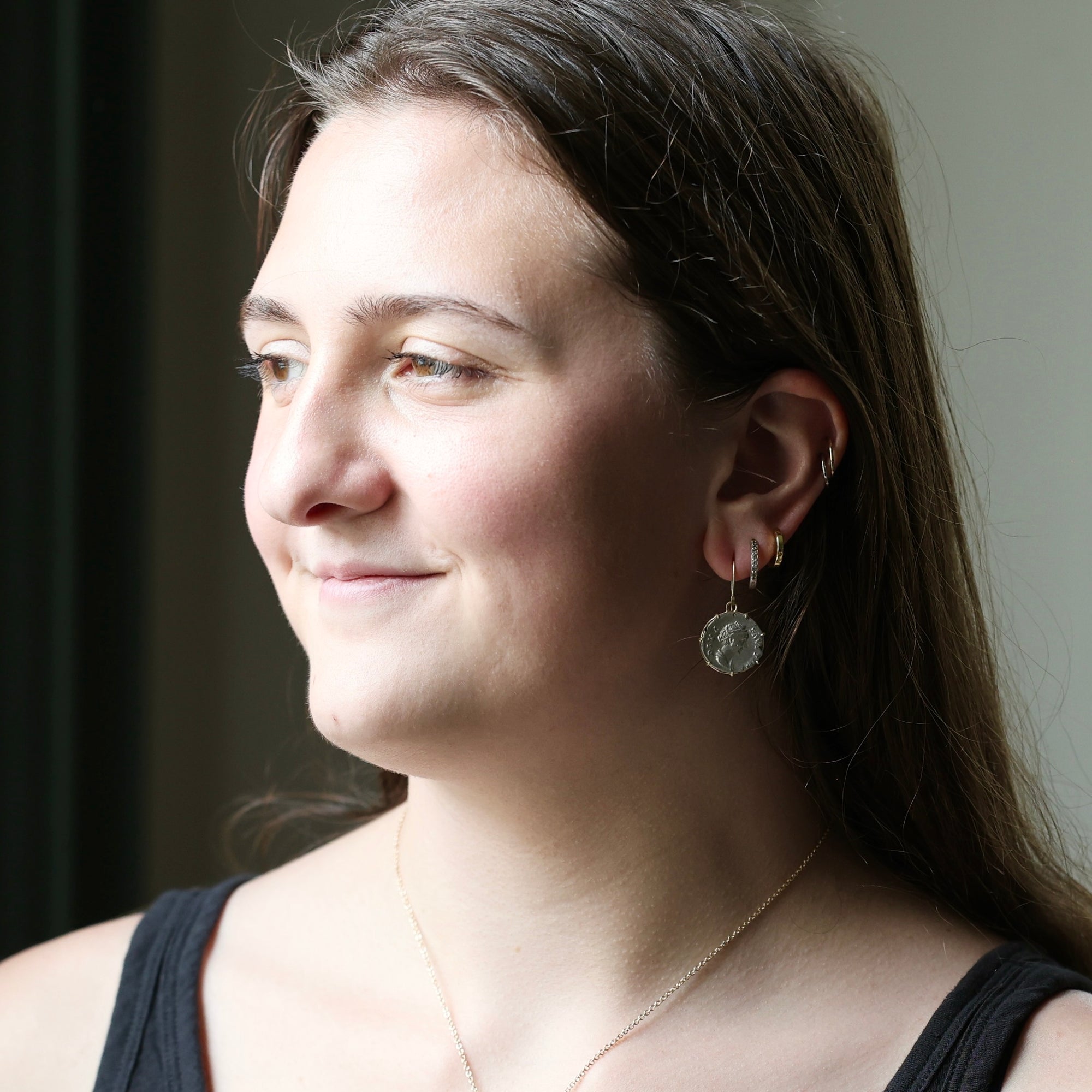 A woman in profile wearing a silver coin pendant earring and a delicate gold chain necklace, shown in a close-up portrait.