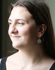 A woman in profile wearing a silver coin pendant earring and a delicate gold chain necklace, shown in a close-up portrait.