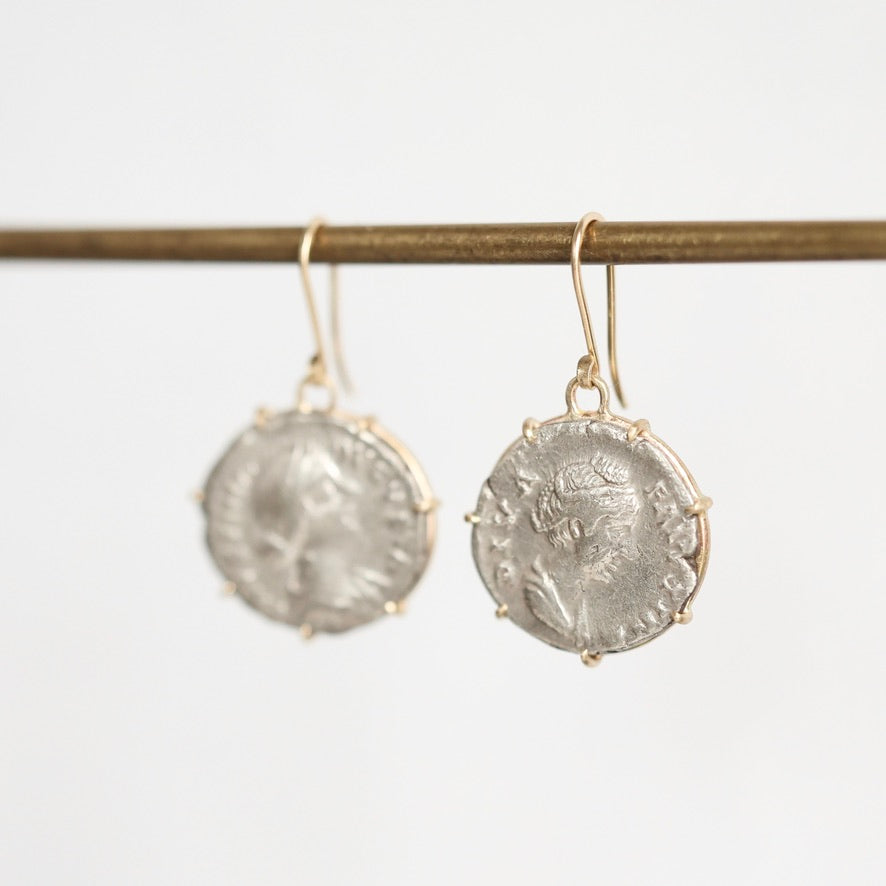 A pair of dangling earrings with round silver-toned coin-style medallions in prong settings and gold-toned hooks, hanging from a horizontal rod against a plain light background.