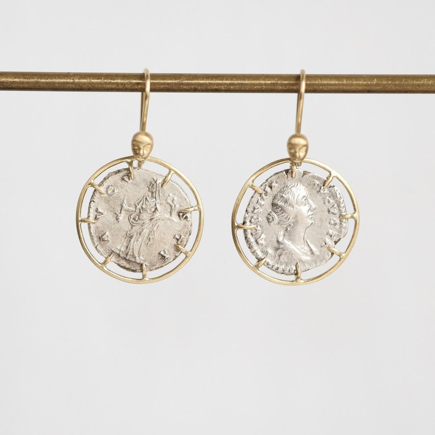 A pair of gold and silver drop earrings featuring circular ancient coin designs, hanging from a gold rod against a light gray background.