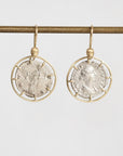 A pair of gold and silver drop earrings featuring circular ancient coin designs, hanging from a gold rod against a light gray background.