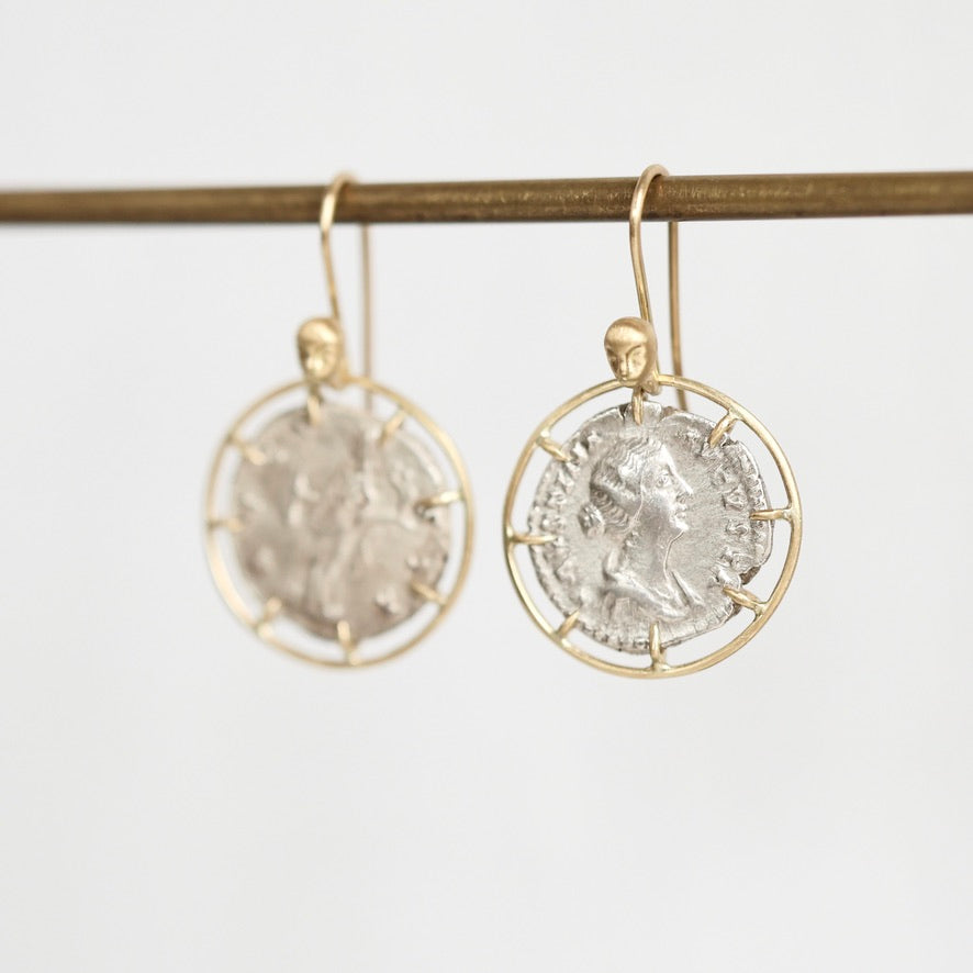 A pair of gold-tone dangle earrings with circular wire frames holding antique silver coins, hanging from a gold rod against a light gray background.
