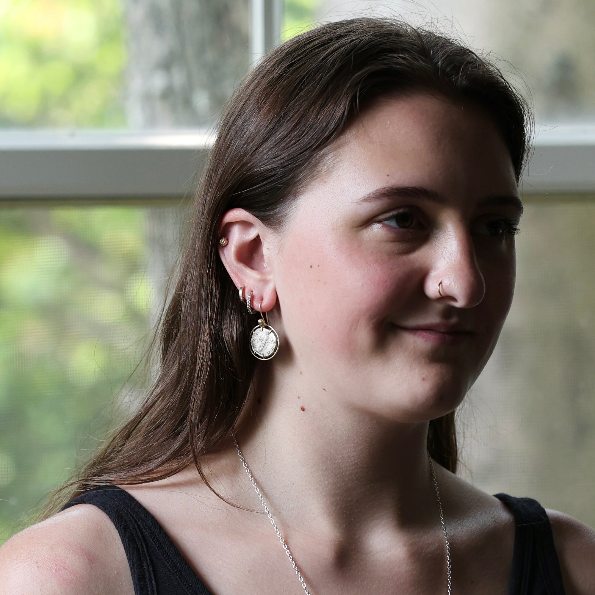 A woman with long brown hair is shown in profile, wearing a silver hoop earring with a white oval stone and a delicate silver necklace. She is wearing a black top, and the background is a softly blurred window with green foliage outside.