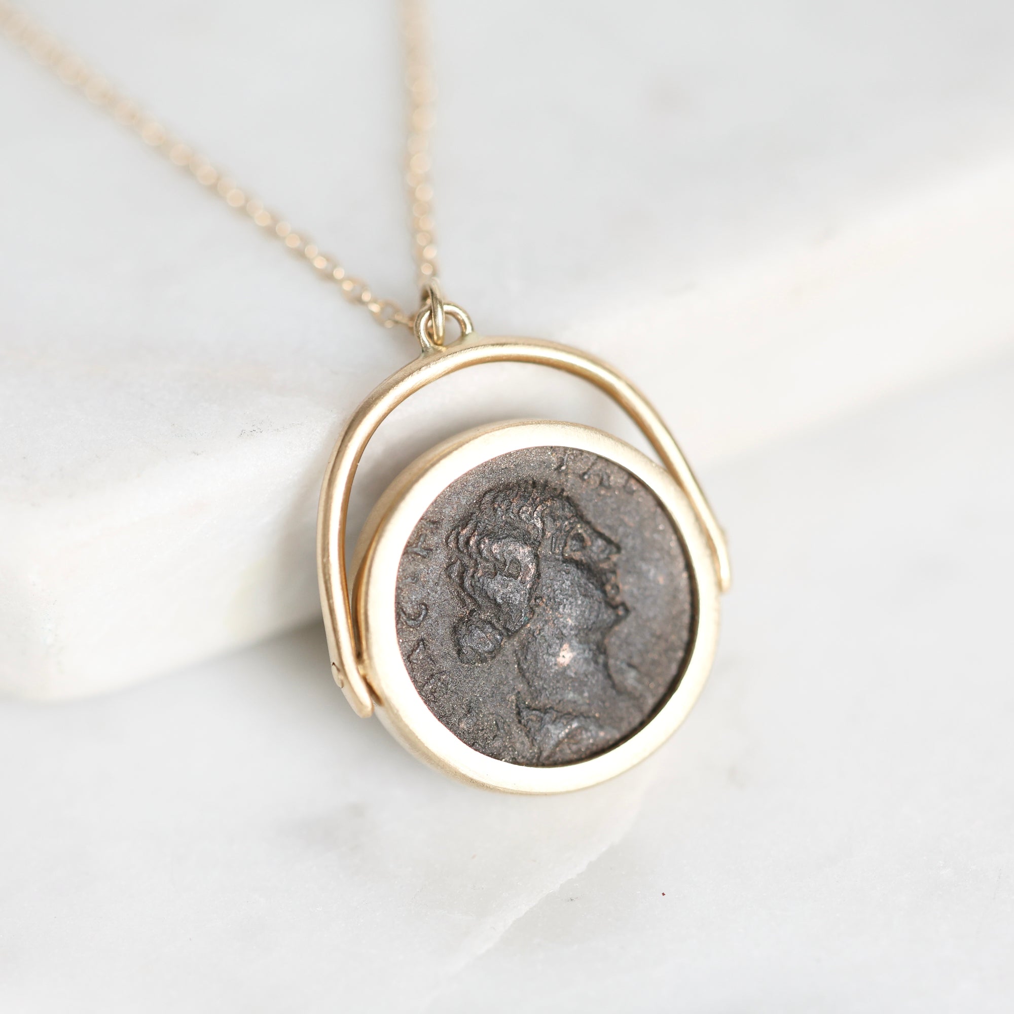 A gold necklace with a circular pendant featuring an antique silver coin profile, displayed on a white marble surface.