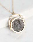 A gold necklace with a circular pendant featuring an antique silver coin profile, displayed on a white marble surface.