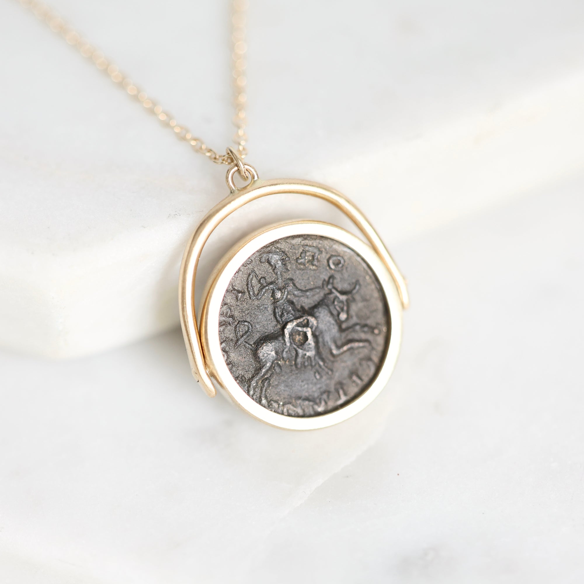 A gold necklace with a circular pendant featuring an antique-style silver coin medallion, displayed on a white marble surface.