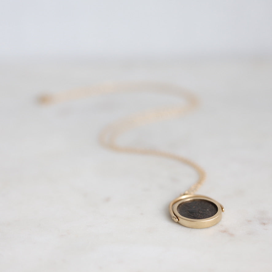 A gold locket necklace with a dark circular medallion, resting on a light marble surface with a soft-focus background.
