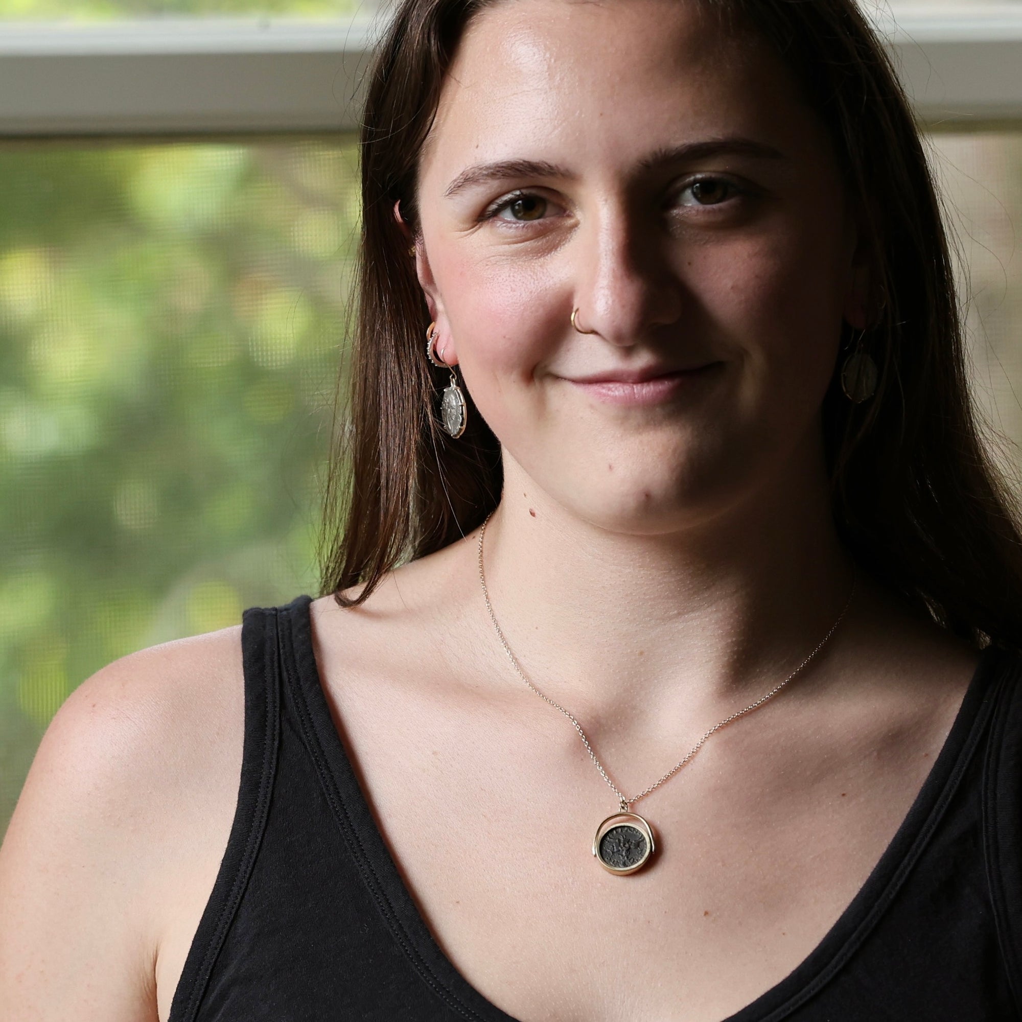 A woman wearing a delicate gold chain necklace with a round dark stone pendant, styled with silver drop earrings and a black tank top, photographed in front of a softly blurred window.