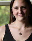 A woman wearing a delicate gold chain necklace with a round dark stone pendant, styled with silver drop earrings and a black tank top, photographed in front of a softly blurred window.