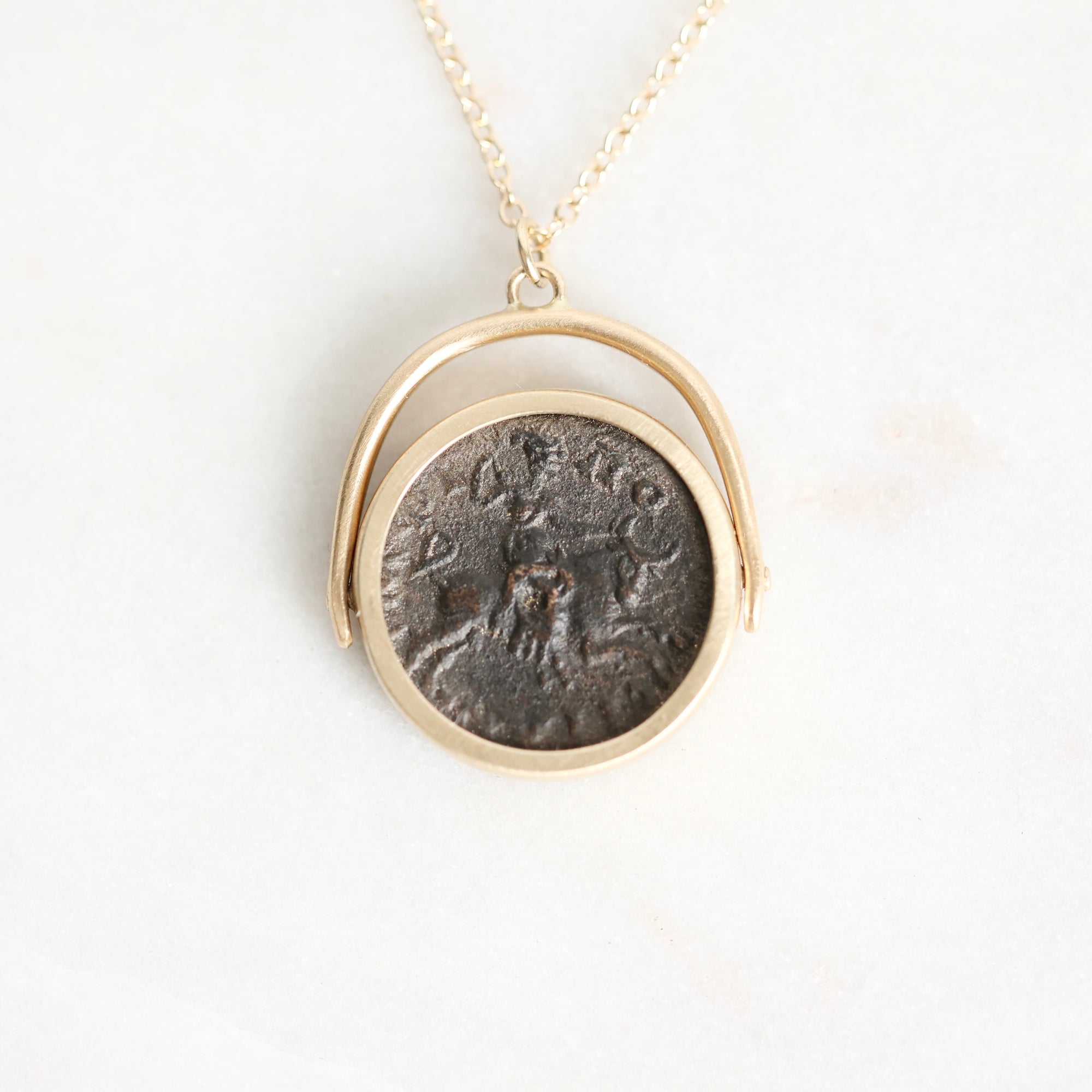 A gold necklace with a circular pendant featuring an antique dark bronze coin, displayed on a clean white background.
