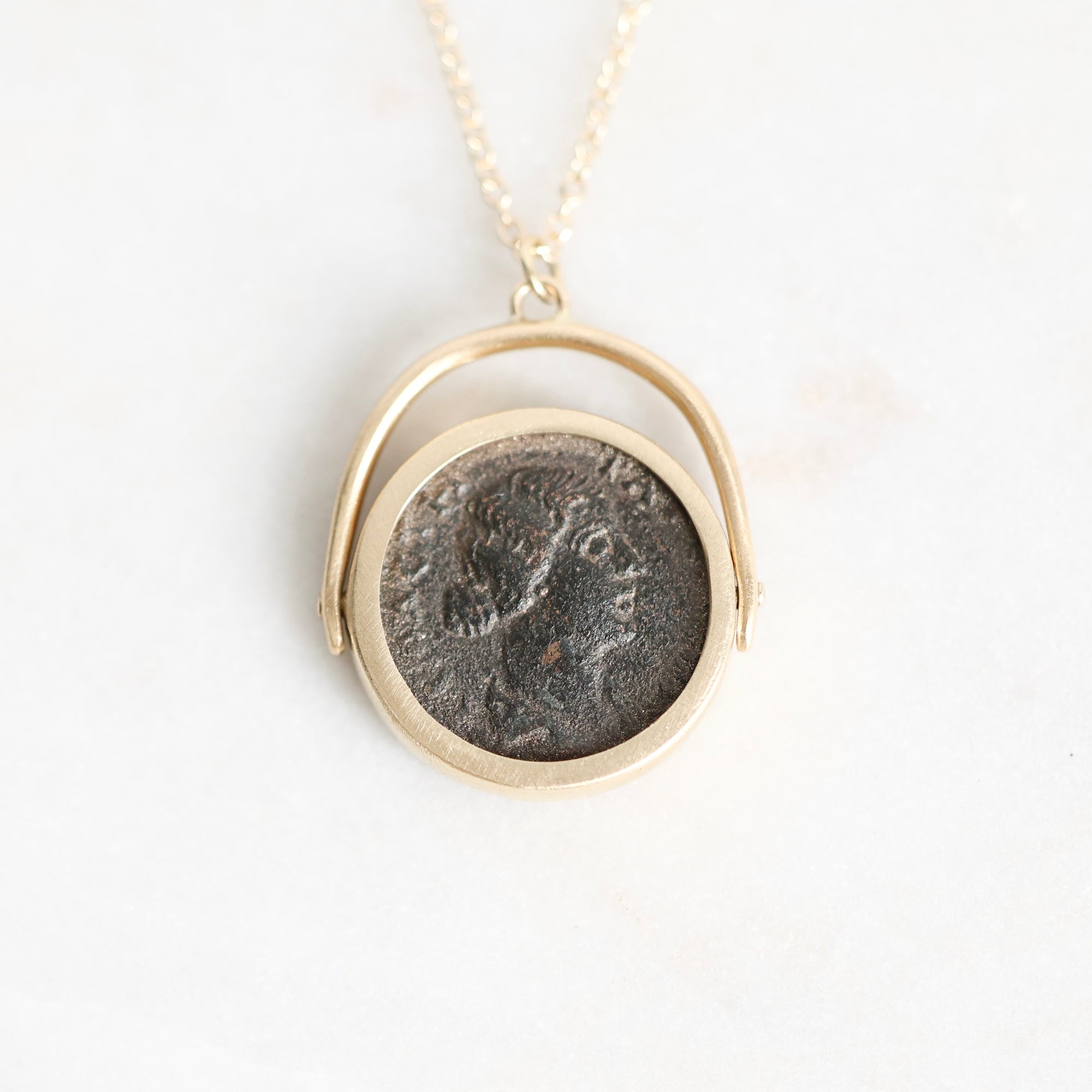 A gold pendant necklace featuring a circular coin charm with an embossed profile portrait, suspended from a delicate gold chain against a plain white background.