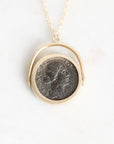 A gold pendant necklace featuring a circular coin charm with an embossed profile portrait, suspended from a delicate gold chain against a plain white background.