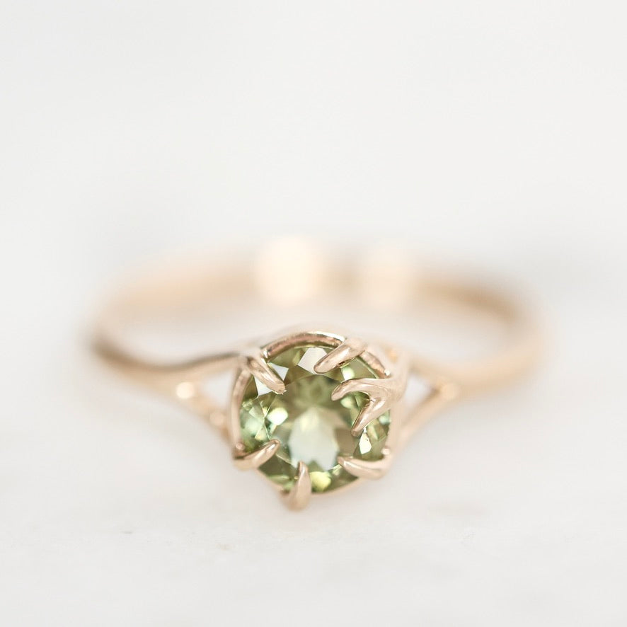 A close-up shot of a rose gold ring featuring a round-cut light green gemstone in a prong setting, set against a soft, out-of-focus white background.