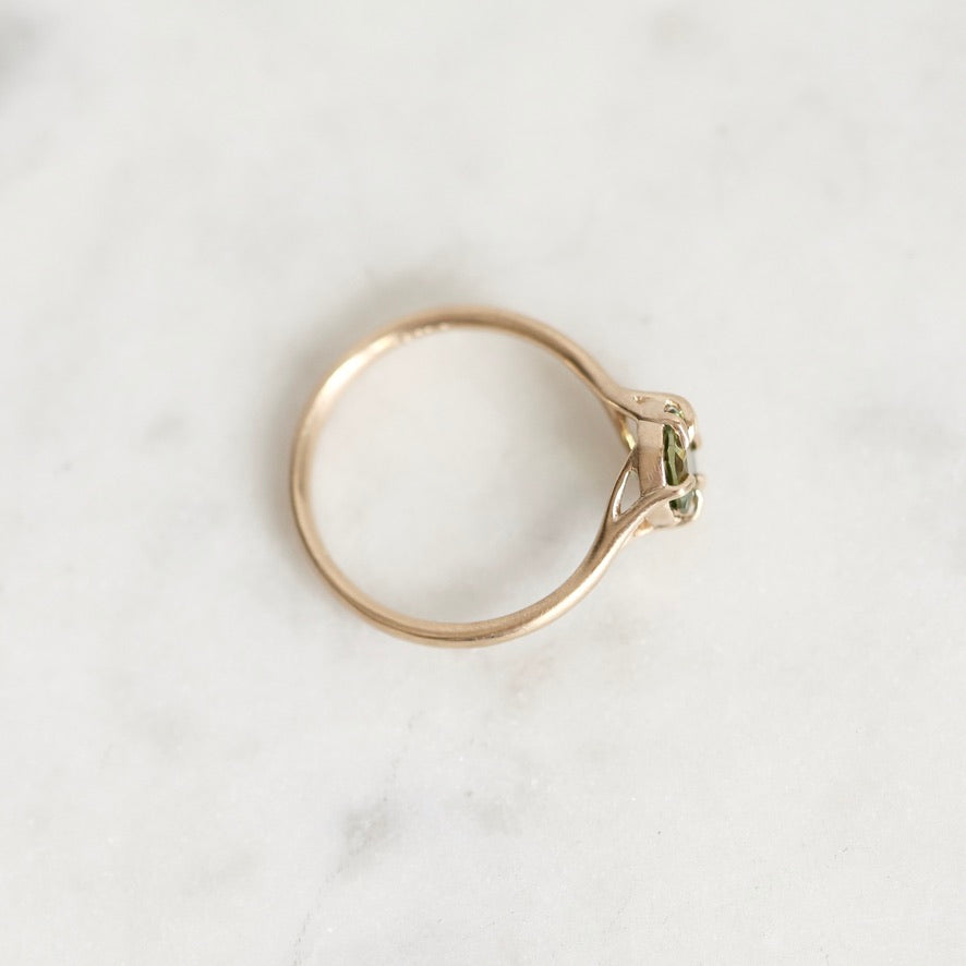 A delicate gold ring with a small green gemstone, photographed from an overhead angle on a light gray marble surface.