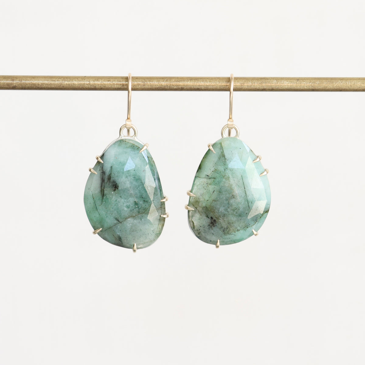 A pair of teardrop-shaped drop earrings with faceted light green and blue marbled gemstones in gold prong settings, hanging from gold hooks on a horizontal gold rod against a plain off-white background.