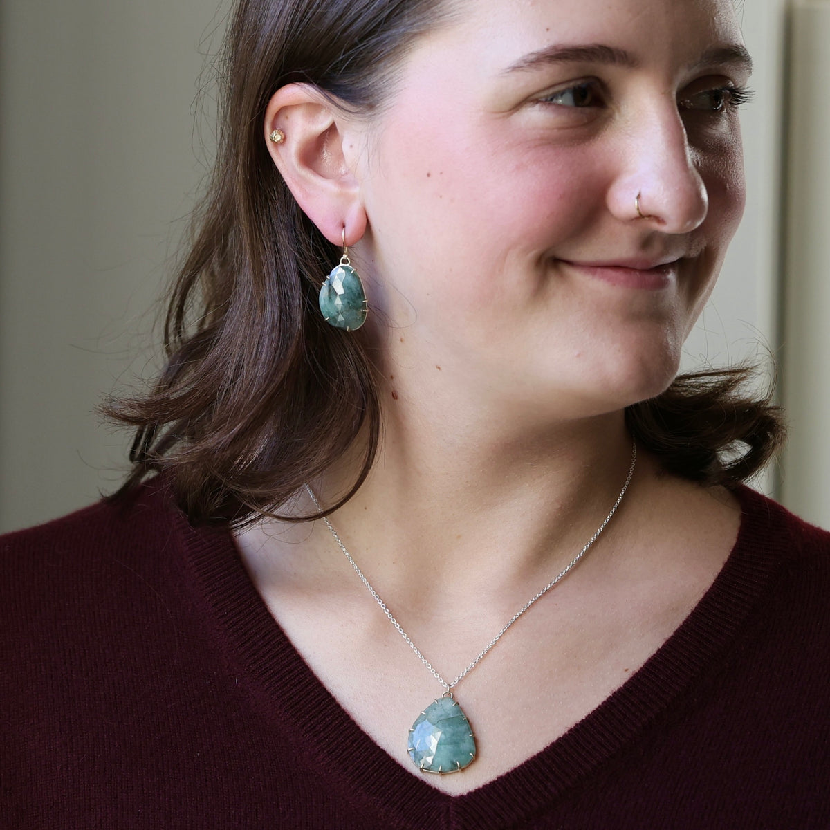 A woman wearing a silver-toned necklace and matching drop earrings with oval light blue-green stones, shown in a close-up portrait against a soft beige background.