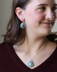 A woman wearing a silver-toned necklace and matching drop earrings with oval light blue-green stones, shown in a close-up portrait against a soft beige background.