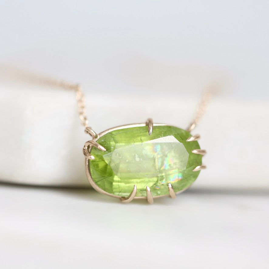 A close-up shot of a gold necklace with an oval-cut green gemstone pendant, resting on a white surface.