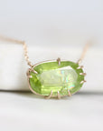A close-up shot of a gold necklace with an oval-cut green gemstone pendant, resting on a white surface.