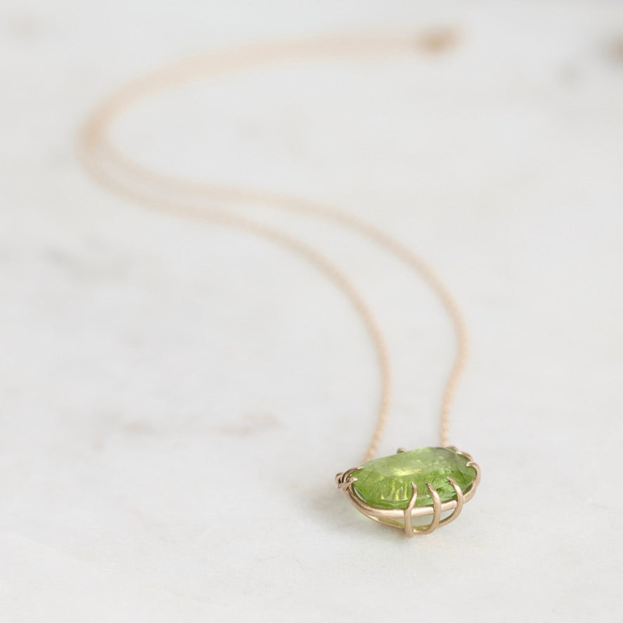 A delicate gold necklace with an oval green gemstone pendant, resting on a light marble surface.