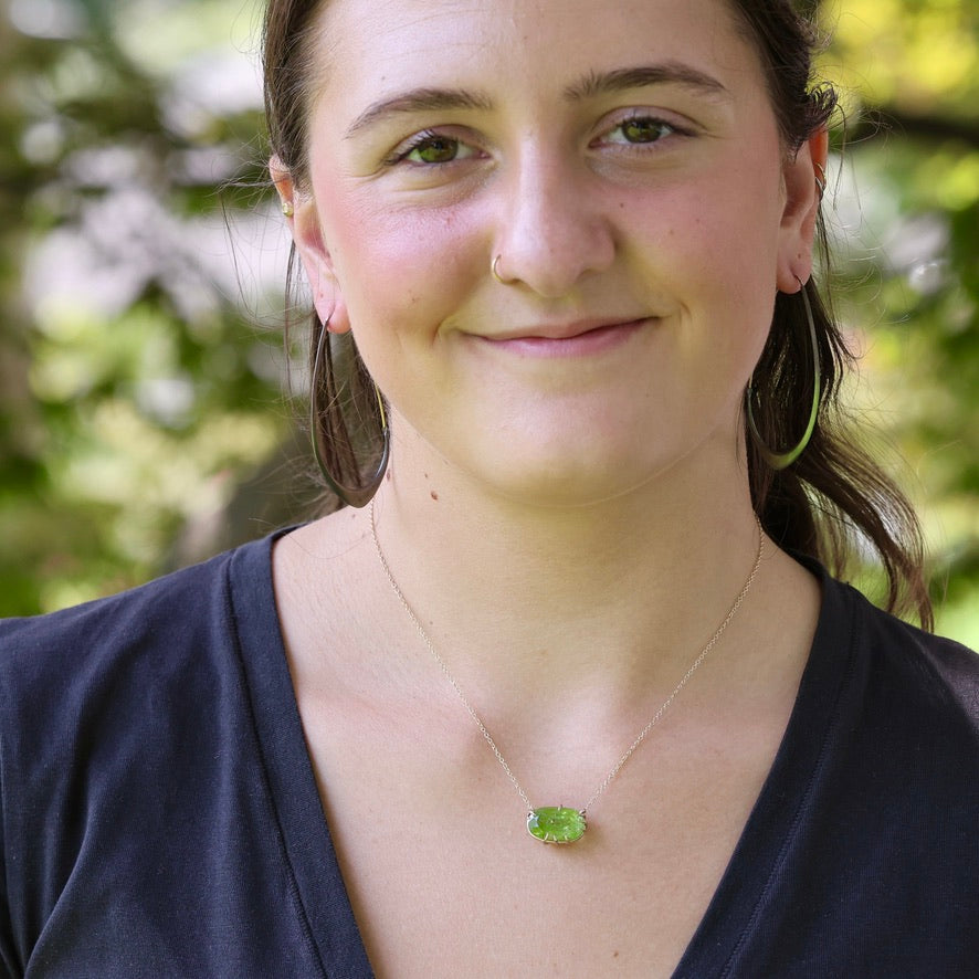 A woman wearing a delicate silver chain necklace with a small, vibrant green oval pendant, photographed outdoors with a soft-focus background of green foliage.