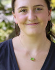 A woman wearing a delicate silver chain necklace with a small, vibrant green oval pendant, photographed outdoors with a soft-focus background of green foliage.