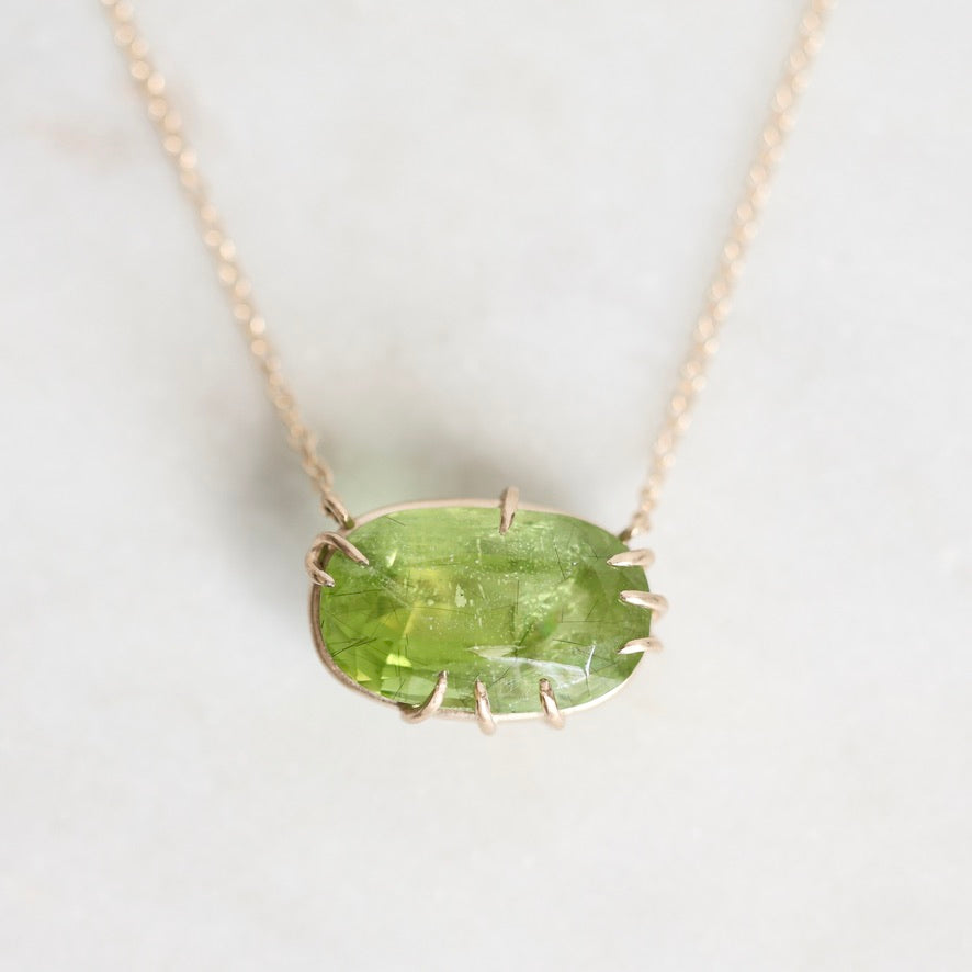 A gold necklace with a bezel-set oval green gemstone pendant, displayed on a light marble surface.