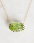 A gold necklace with a bezel-set oval green gemstone pendant, displayed on a light marble surface.