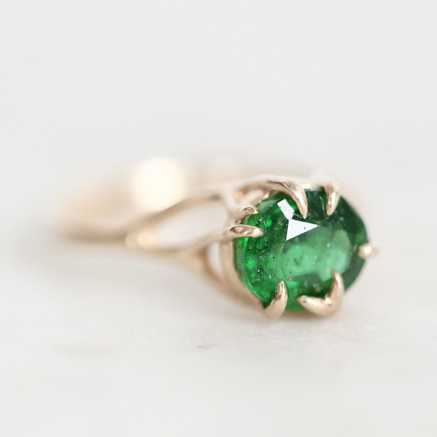 A close-up shot of a gold ring featuring a round-cut green emerald gemstone in a prong setting, resting on a light, neutral surface.