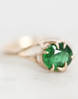 A close-up shot of a gold ring featuring a round-cut green emerald gemstone in a prong setting, resting on a light, neutral surface.