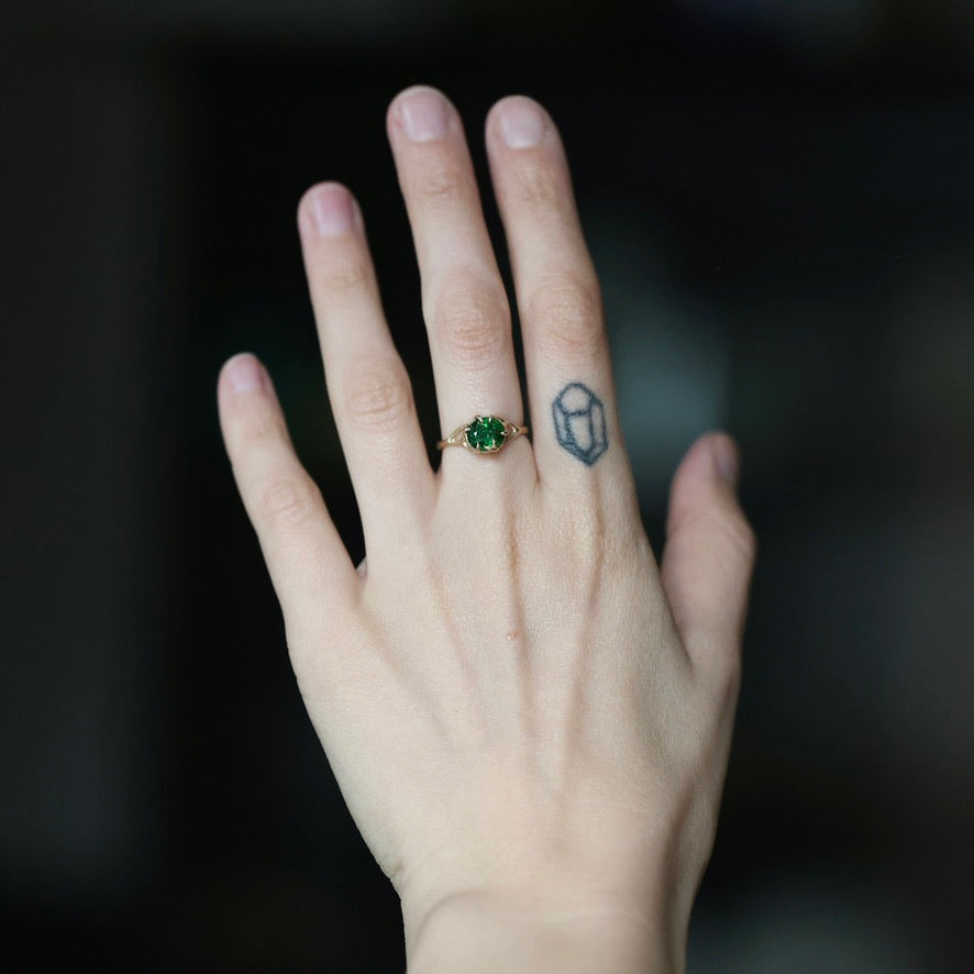 A close-up of a person's hand wearing a small gold ring with a green gemstone, with a geometric tattoo on the middle finger, against a dark blurred background.
