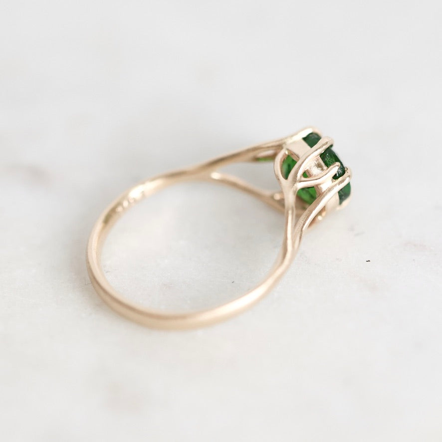 A gold ring with a green gemstone in a prong setting, photographed on a light marble surface.