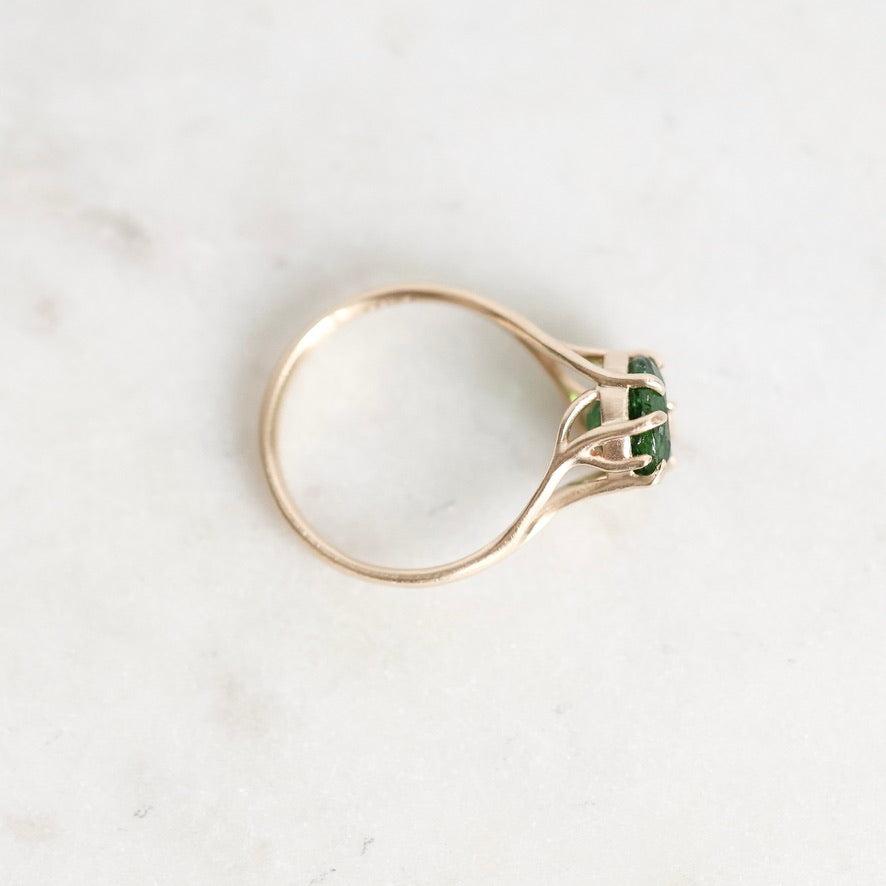 A gold ring with a split shank and a small green rectangular gemstone, photographed on a light marble surface.