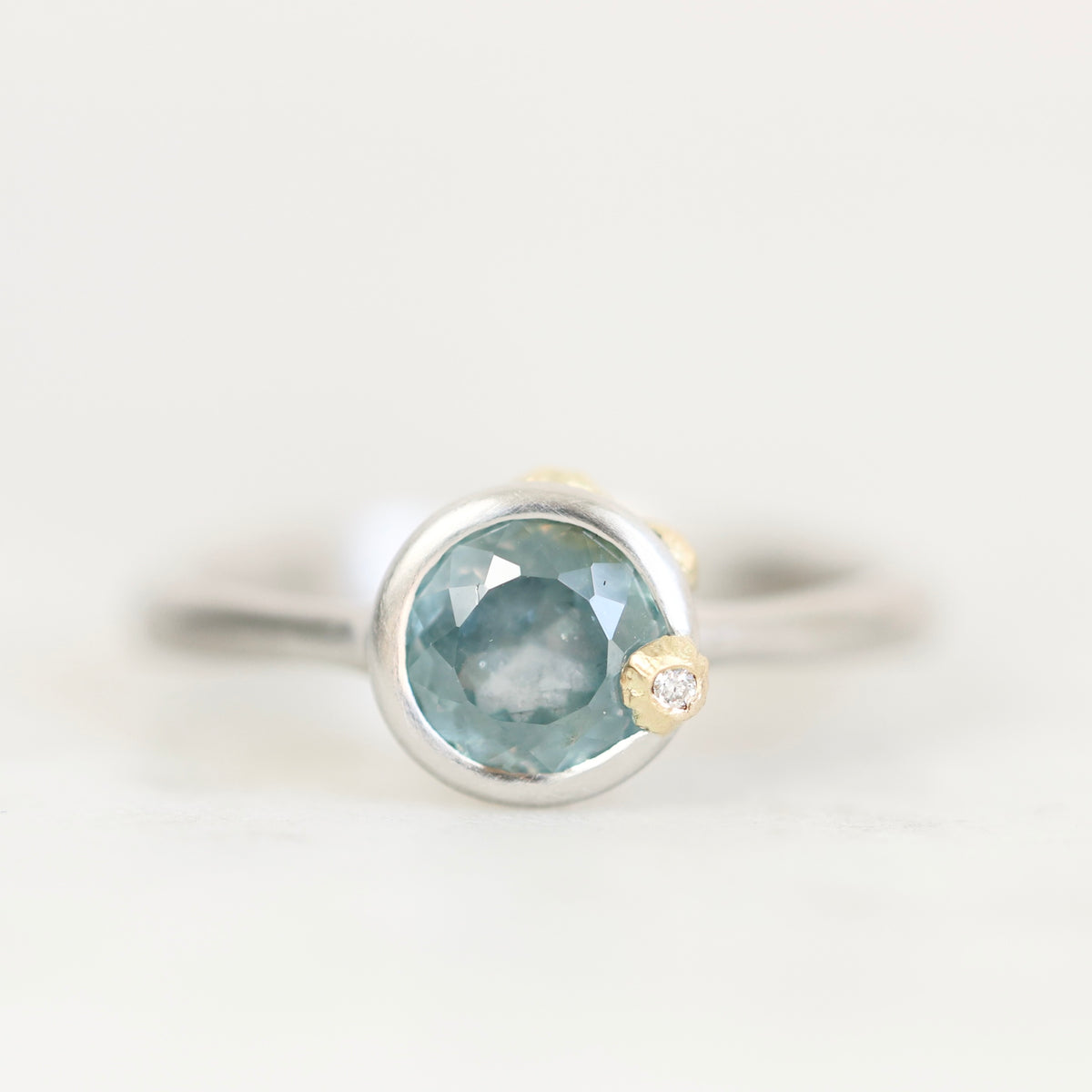 A silver ring with a round faceted light blue gemstone and a small gold accent on the band, photographed against a clean white background.
