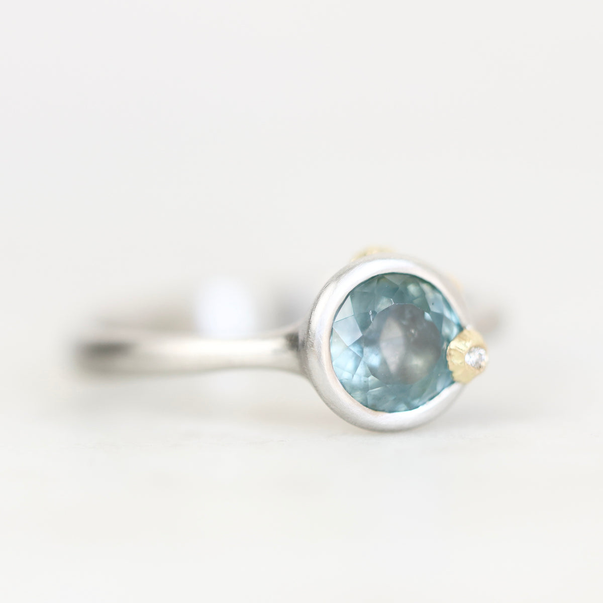 A silver ring featuring a round light blue gemstone and a small gold accent on the band, photographed against a soft white background.