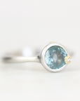 A silver ring featuring a round light blue gemstone and a small gold accent on the band, photographed against a soft white background.